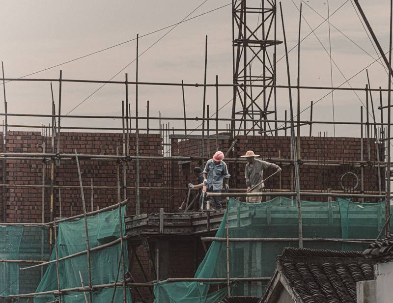 Two builders on scaffolding working on a brick wall under a cloudy sky